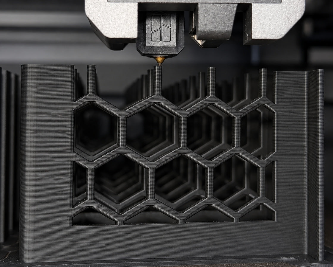 Close-up of a 3D printer nozzle building the honeycomb wall of a black Slabster riser, layer lines visible